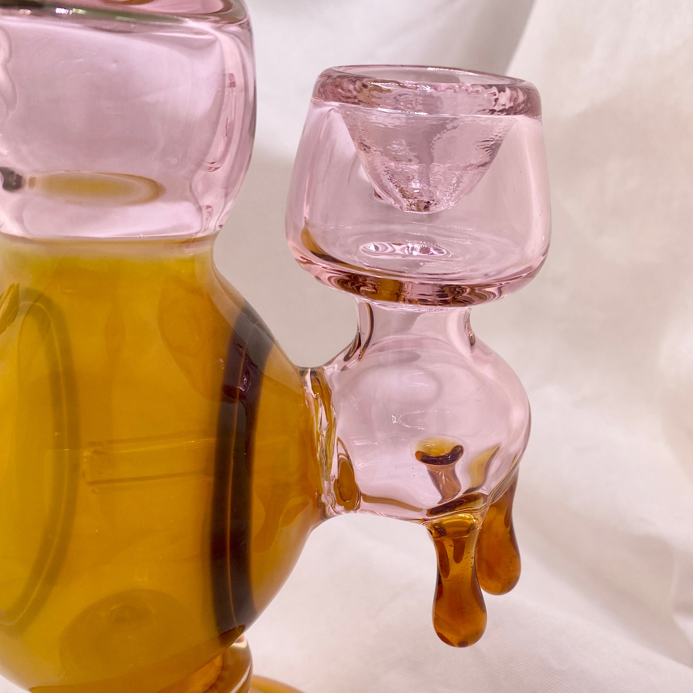 Pink glass container with a spout pouring amber liquid onto a white surface.